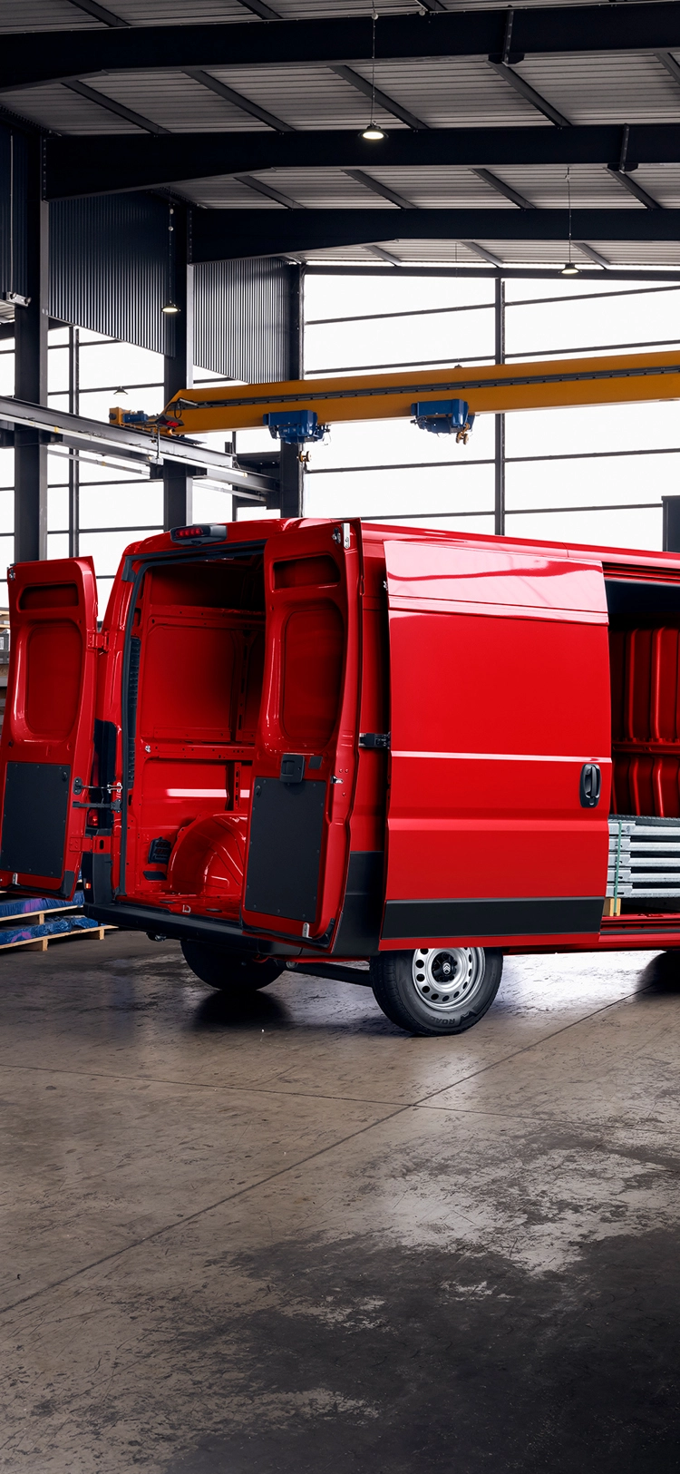 Widok 3/4 z tyłu czerwonego Citroëna Jumper z otwartymi drzwiami przedziału ładunkowego stojącego w hali magazynowej, ilustrujący obszerną przestrzeń ładunkową, wytrzymałą konstrukcję i wszechstronność zastosowań w firmie.