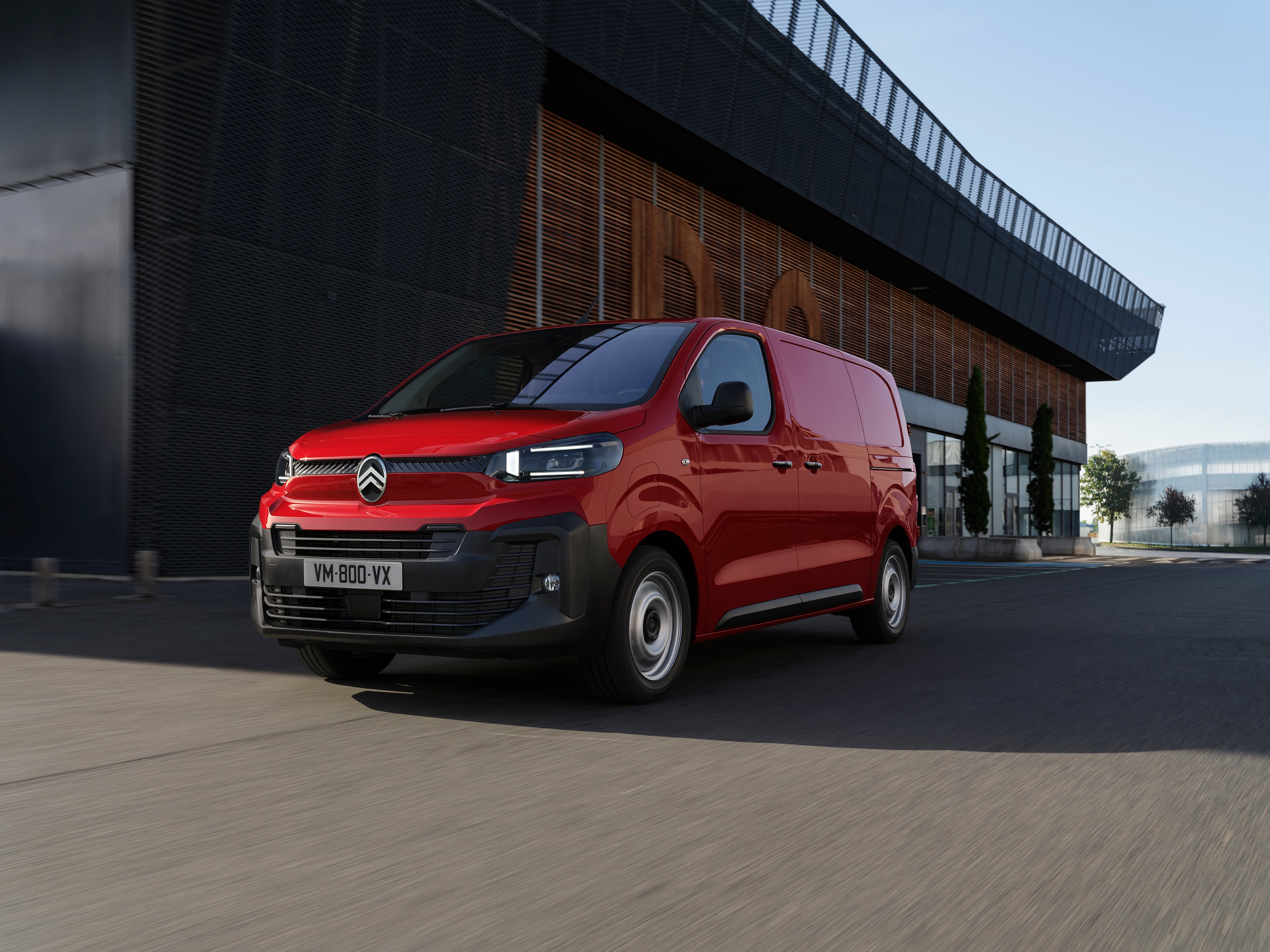 Widok 3/4 z przodu czerwonego Citroëna Jumpy zaparkowanego przy nowoczesnym budynku, pokazujący profesjonalny design, praktyczność oraz wyrazistą, pewną sylwetkę.