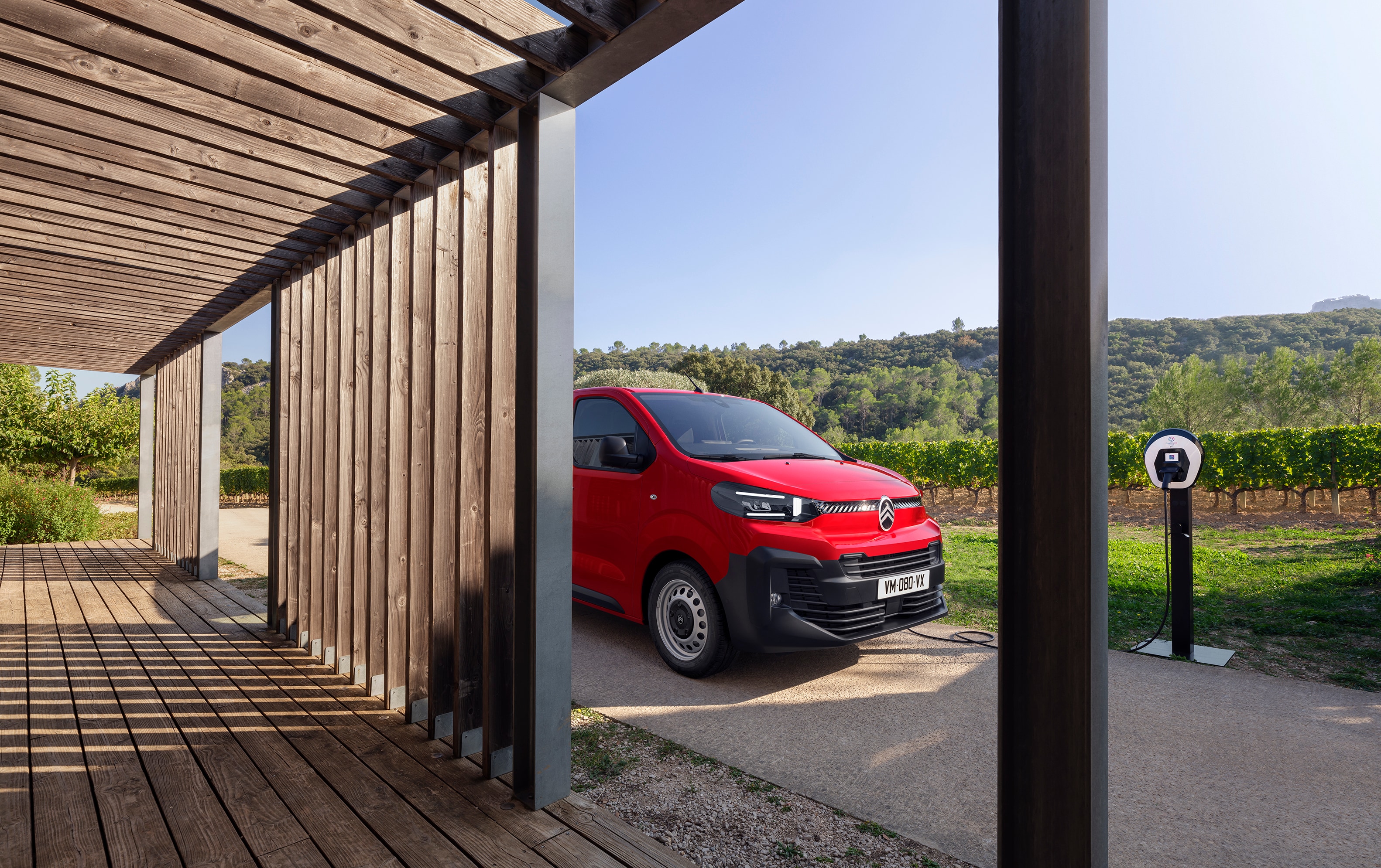 Widok 3/4 z boku czerwonego Citroëna ë‑Jumpy zaparkowanego przed drewnianym budynkiem, pokazujący nowoczesny elektryczny design, codzienną funkcjonalność i pewną, profesjonalną prezencję.