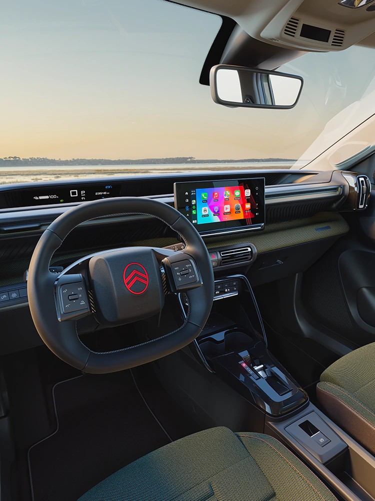 Widok z miejsca kierowcy na deskę rozdzielczą Citroëna ë-C3 Outdoor z ekranem dotykowym, ukazujący łączność i komfort Advanced Comfort podczas podróży kompaktowym SUV-em